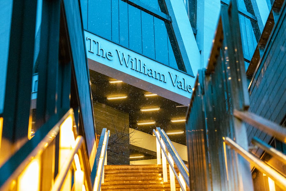 Photograph of the entrance to The William Vale hotel at dusk, taken from a low angle looking upward. The exterior features modern architectural design with blue panels, illuminated by warm, yellow lighting. Metal railings and wooden steps lead up to the building's doorway, where a large sign displays the hotel name. The surroundings include part of a metal staircase or framework on the right side, and the scene is illuminated by external lighting, highlighting the building's materials and structure. This image captures an urban hotel environment suitable for house removals or moving services that involve hotel relocations or accommodation changes, with Maida Vale Removals potentially assisting with such tasks.