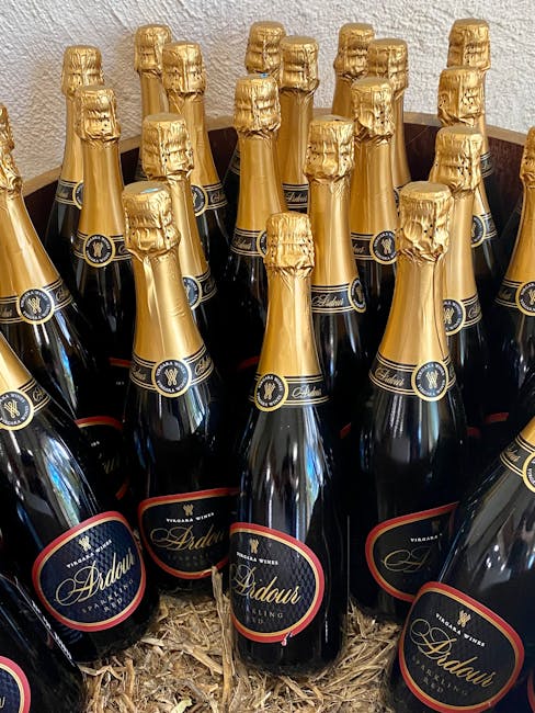 Multiple bottles of Ardau sparkling wine with gold foil wrapping on the necks and black and red labels, arranged upright in a cardboard box filled with straw-like packing material. The bottles are positioned closely together, occupying the entire frame, with some bottles overlapping each other. The background features a textured beige wall, and the bottles are placed on a wooden surface. This image depicts the careful packing of beverages, likely during a home relocation or moving process, such as those managed by Maida Vale Removals, emphasizing the importance of secure packing for fragile items during furniture transport and home removals.