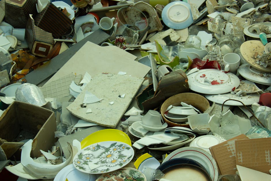 A large pile of broken and discarded household ceramic dishes, cups, plates, and bowls, mixed with pieces of broken glass, plastic, and cardboard packaging. Some items, such as a yellow plastic tray and a floral-patterned plate, are more intact, while others are shattered or severely damaged. The debris is spread across a cluttered indoor or outdoor space, with parts of a wooden box, a granite countertop fragment, and a grey tiled surface visible beneath the mess. The scene suggests a collection of refuse or discarded items, potentially prepared for disposal or recycling as part of a house clearance or relocation process. The image exemplifies the typical clutter involved in household removal services, such as those provided by Maida Vale Removals, which handle furniture and waste removal in preparation for home relocation.