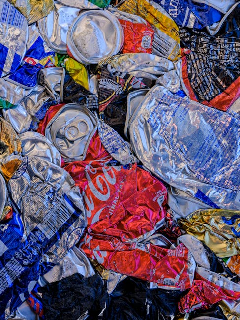 A close-up view of crushed, flattened aluminium beverage cans, primarily in silver, with some featuring remnants of labels in red, blue, and yellow. The cans are piled together, with some showing visible pull-tabs and indentations, indicating they have been emptied and compressed for recycling or disposal. The background is filled with more flattened cans, creating a textured, cluttered appearance. This image illustrates waste material likely prepared for collection or recycling as part of a house removal or waste disposal process. The setting appears to be indoors, with no clear indication of a specific environment, but the focus is on the discarded cans, suggesting a step in packing or clearing during a home relocation or waste management activity. Maida Vale Removals offers services related to moving and disposal, including handling various household waste like empty cans.