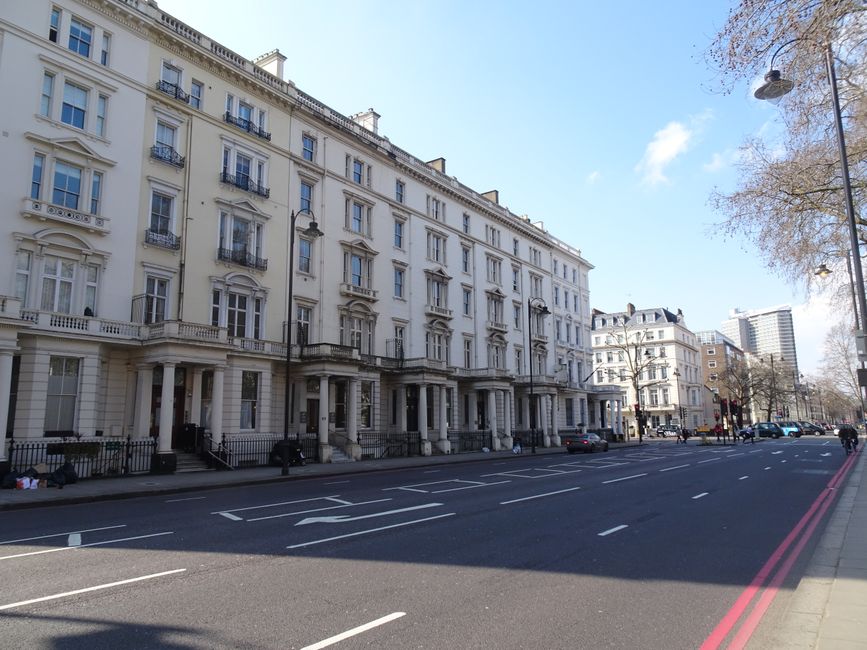 A daytime street scene showcasing a multi-storey brick building with large arched and rectangular windows, situated adjacent to other historic and modern buildings in an urban area. In front of the bu