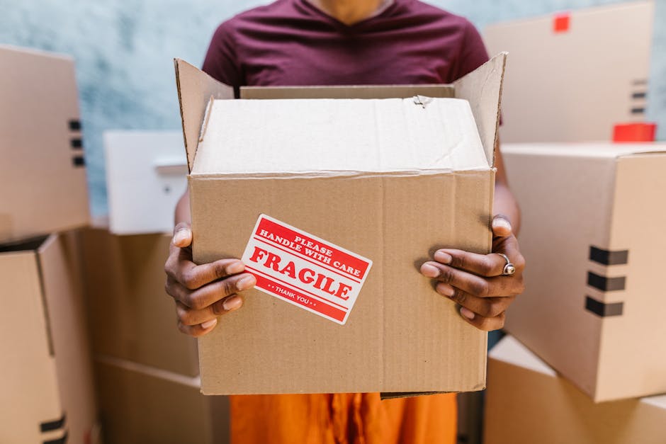 A person holding an open cardboard box with a red and white 'FRAGILE' handling sticker, surrounded by several packed boxes in a warehouse or storage area. The individual, wearing a maroon shirt, grips the box from the sides, with their hands visible displaying a wedding ring on one finger. The background contains other closed cardboard boxes stacked on top of each other in a well-lit environment, indicating a packing or home relocation process, possibly related to house removals or furniture transport services provided by Maida Vale Removals in the context of Clifton Road moves.