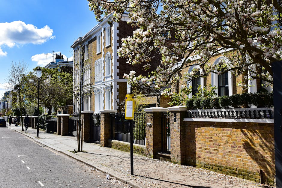 The image depicts a residential street scene featuring a row of Victorian-style terraced houses with brick facades and white window frames, situated behind a brick and wrought iron fence with decorative railings. Several trees with white blossoms line the sidewalk, casting shadows on the pavement, which is paved with concrete slabs. A yellow bus stop sign is affixed to a black pole near the sidewalk, adjacent to a young, leafless tree. The sky above is clear with a few scattered clouds, and sunlight illuminates the scene, highlighting the structure and greenery. This context aligns with house removals and home relocation processes, as Maida Vale Removals regularly assists with moving services in this area, encompassing packing, transportation, and logistics for residential properties.