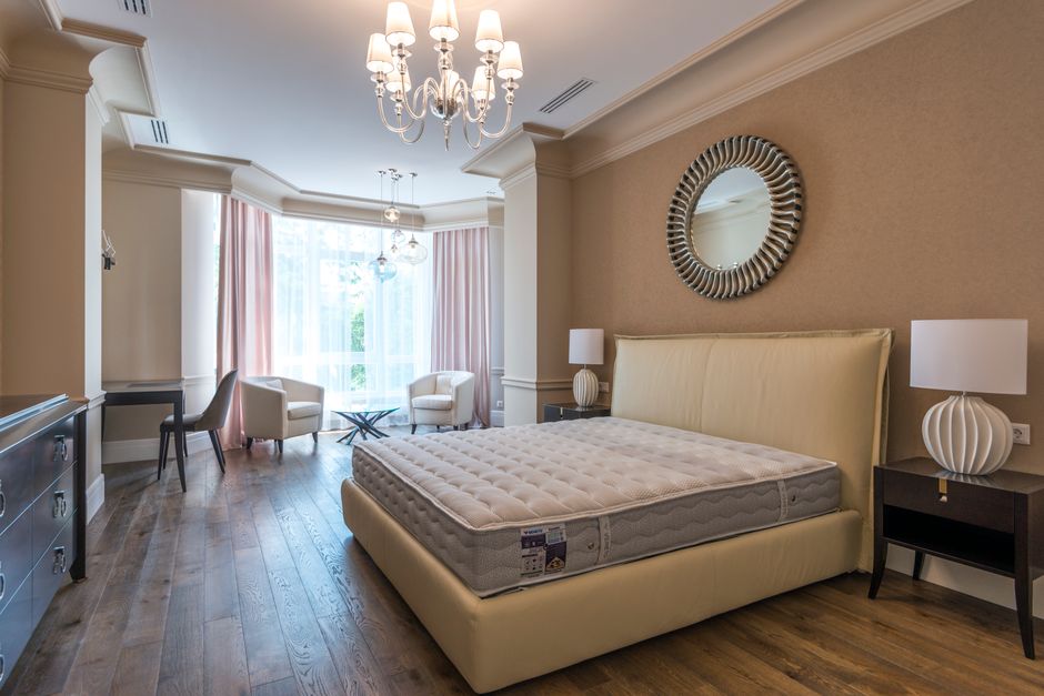A spacious bedroom with beige walls and a wooden floor, featuring a large cream upholstered bed with a mattress still in packaging, positioned against the right wall. On each side of the bed, there are black bedside tables topped with white, sculptural table lamps. Above the headboard, a round decorative mirror with a textured frame is mounted on the wall. To the left of the bed, there is a tall white curtain covering a wide window that allows natural light into the room, with pink curtains framing the window. The window opens onto a small balcony or patio with a view of green foliage. Near the window, there are seating arrangements comprising two cream armchairs, a small glass-top table between them, and a black side table with a chair nearby, indicative of a space prepared for moving or packing activities. The ceiling features a decorative chandelier with multiple light bulbs, and additional lighting is provided by a smaller chandelier or ceiling light. Various packing supplies, including cardboard boxes and plastic wrap, are visible on the floor near the bed, consistent with a preparatory phase for house removals. The overall setting appears clean, well-lit, and in the process of a home relocation, with visible furniture and packing materials revealing the moving process supported by Maida Vale Removals.
