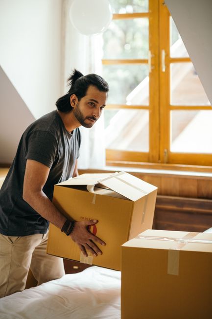 A man with dark hair and a beard, dressed in a grey t-shirt, is indoors during a home relocation, holding a cardboard box with red handles while preparing to move it. The room has natural light coming through a large wooden-framed window with a view of greenery outside. Partially visible white curtains hang beside the window, and a white spherical ceiling light is in the upper left corner. The man is standing on a bed with a white duvet, surrounded by several other packed boxes, some with packing tape. The wooden floor and wall paneling suggest a cozy, residential interior. This scene illustrates the packing and loading process involved in house removals, with Maida Vale Removals ensuring careful handling of furniture and boxes for an efficient move.