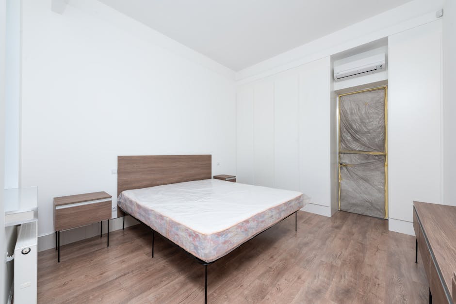A spacious bedroom with beige walls and a wooden floor, featuring a large cream upholstered bed with a mattress still in packaging, positioned against the right wall. On each side of the bed, there are black bedside tables topped with white, sculptural table lamps. Above the headboard, a round decorative mirror with a textured frame is mounted on the wall. To the left of the bed, there is a tall white curtain covering a wide window that allows natural light into the room, with pink curtains framing the window. The window opens onto a small balcony or patio with a view of green foliage. Near the window, there are seating arrangements comprising two cream armchairs, a small glass-top table between them, and a black side table with a chair nearby, indicative of a space prepared for moving or packing activities. The ceiling features a decorative chandelier with multiple light bulbs, and additional lighting is provided by a smaller chandelier or ceiling light. Various packing supplies, including cardboard boxes and plastic wrap, are visible on the floor near the bed, consistent with a preparatory phase for house removals. The overall setting appears clean, well-lit, and in the process of a home relocation, with visible furniture and packing materials revealing the moving process supported by Maida Vale Removals.