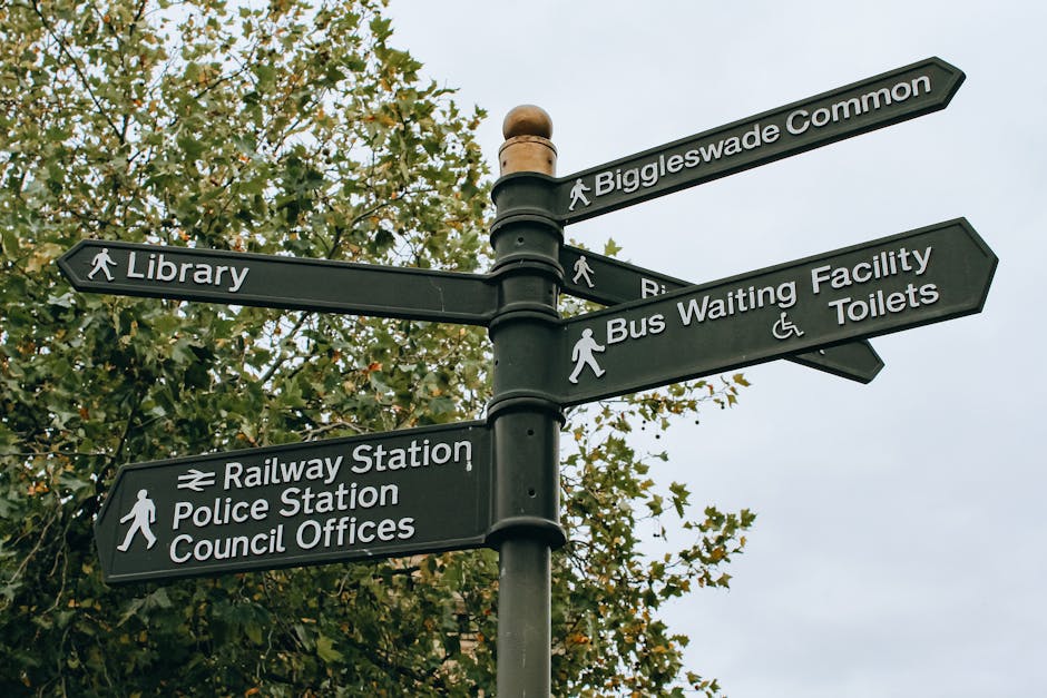 A black multi-directional signpost situated outdoors against a backdrop of leafy trees and an overcast sky, displaying numerous white text panels with directional arrows. The signs point to local landmarks and amenities including Biggleswade Common, Library, Railway Station, Police Station, Council Offices, Bus Waiting Facility, and Toilets, with some signs featuring pedestrian icons. The signpost is part of a street or public area where moving and relocation services by Maida Vale Removals may be relevant, especially in relation to local logistical considerations like parking or directional guidance for home relocations within Westminster and Maida Vale. The surroundings suggest a well-maintained public space used by pedestrians, with no vehicles or moving equipment visible in the image.
