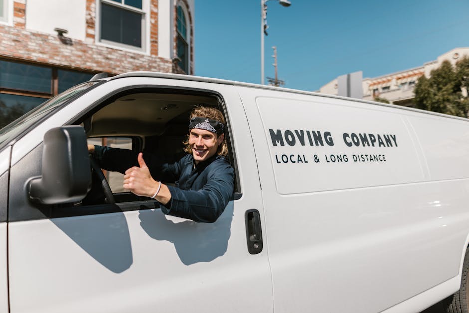 A smiling young man with long hair, wearing a bandana and dark clothing, is sitting in the driver's seat of a white moving van with the signage 'MOVING COMPANY LOCAL & LONG DISTANCE' on the side. He is giving a thumbs-up gesture through the open window. The van is parked outside a residential building in daytime, with clear blue skies overhead. The background shows parts of brick and glass facades, nearby trees, and utility poles, indicating an urban environment. The scene captures the loading or unloading process characteristic of house removals, with the van positioned close to the property for efficient furniture transport and packing during a home relocation, as managed by Maida Vale Removals.