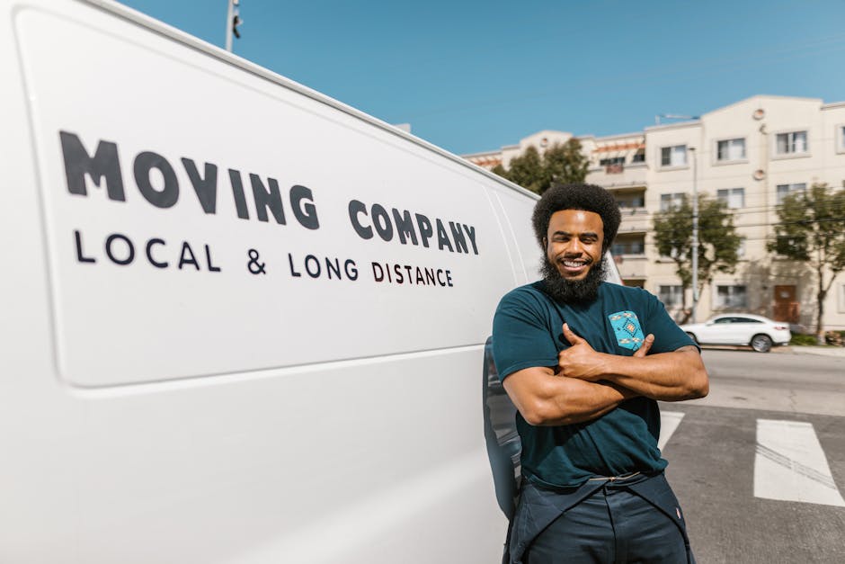 A smiling male mover with a full beard and afro hairstyle stands outside a white moving van with the words 'MOVING COMPANY LOCAL & LONG DISTANCE' printed on its side, near a residential street lined with white multi-storey buildings and parked cars. The mover is wearing a navy blue t-shirt with a small logo on the chest, dark pants, and has his arms crossed. The van is parked on a paved area, partially in front of a pedestrian crossing, with clear blue skies overhead. This scene depicts the home relocation process associated with Maida Vale Removals, highlighting furniture transport and packing and moving logistics typical in house removals within the Maida Vale Warwick Avenue area.