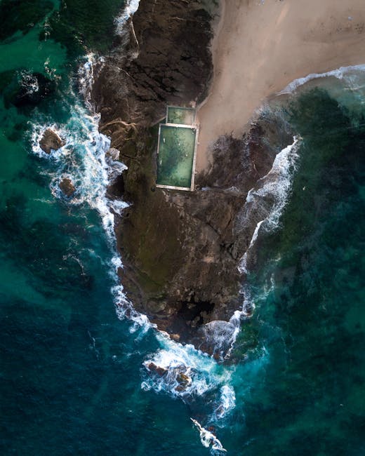 An aerial view of a rocky coastline featuring a small, rectangular swimming pool filled with greenish water, situated on a narrow strip of land between the ocean and a sandy beach. The pool is surrounded by dark rocks and a concrete or paved area, with waves crashing against the rocks and shoreline. A section of the sandy beach extends along the top edge of the image, and the surrounding seawater displays varying shades of blue and turquoise. The scene captures a scenic coastal environment, with the pool partially embedded into the rocky terrain, and no other objects or people visible in the image.