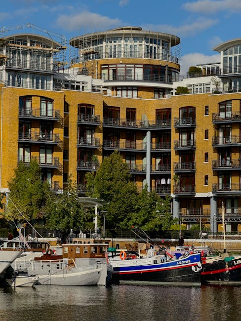 A view of a modern multi-storey residential building with yellow brick exterior and numerous balconies, situated by a river. In the foreground, several boats are moored along the water's edge, including one boat with the name 'Leonid' painted on its side. The scene is captured during daylight with clear blue skies and some scattered clouds. Green trees line the riverbank, adding greenery to the urban landscape. The image appears to illustrate a location relevant to house removals or furniture transport, potentially related to the Maida Vale area, where professional moving services, such as those provided by Maida Vale Removals, operate.